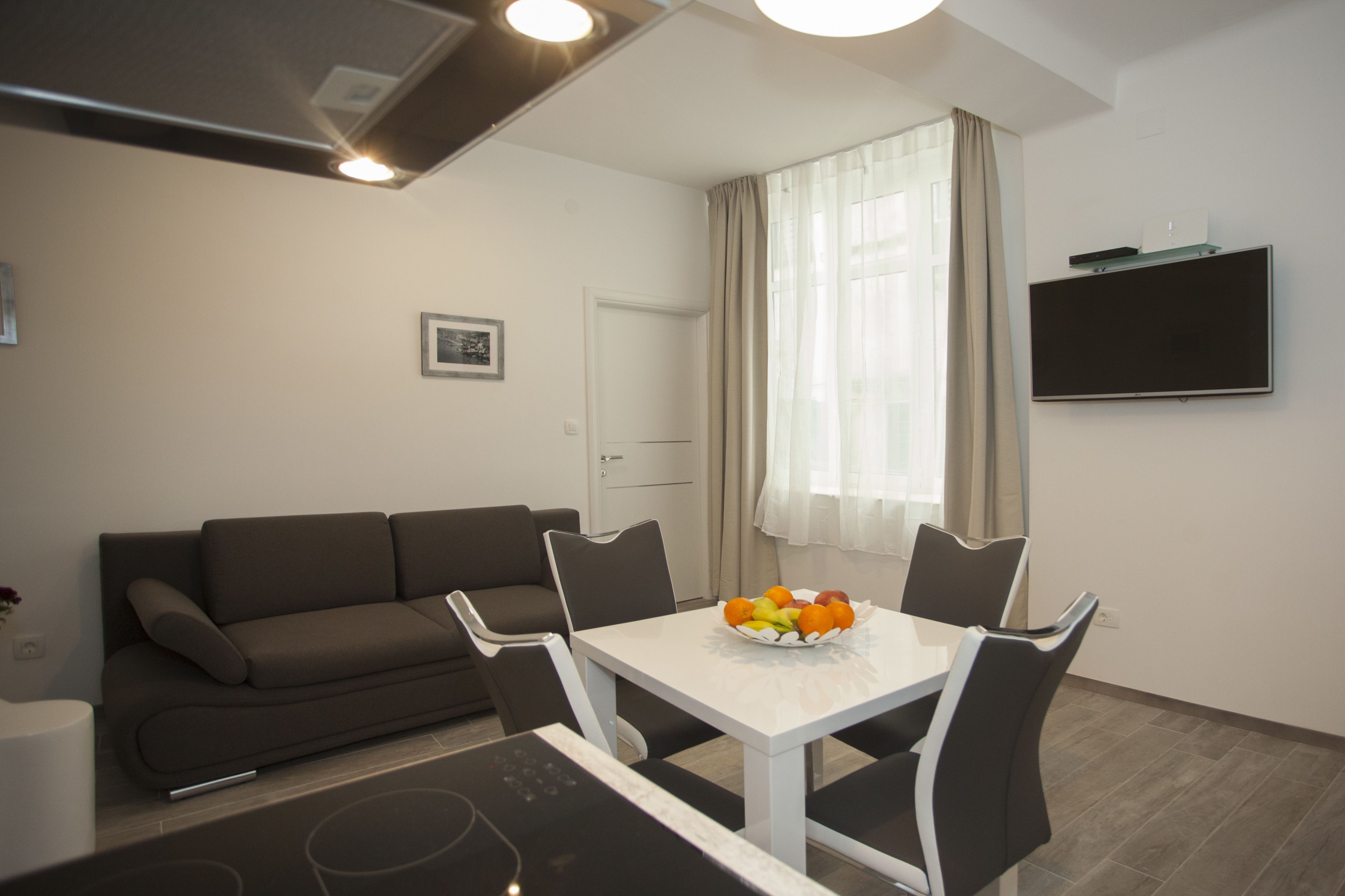 A modern living room with a sofa, dining table, and chairs. There is a flat-screen TV mounted on the wall and a window with curtains.