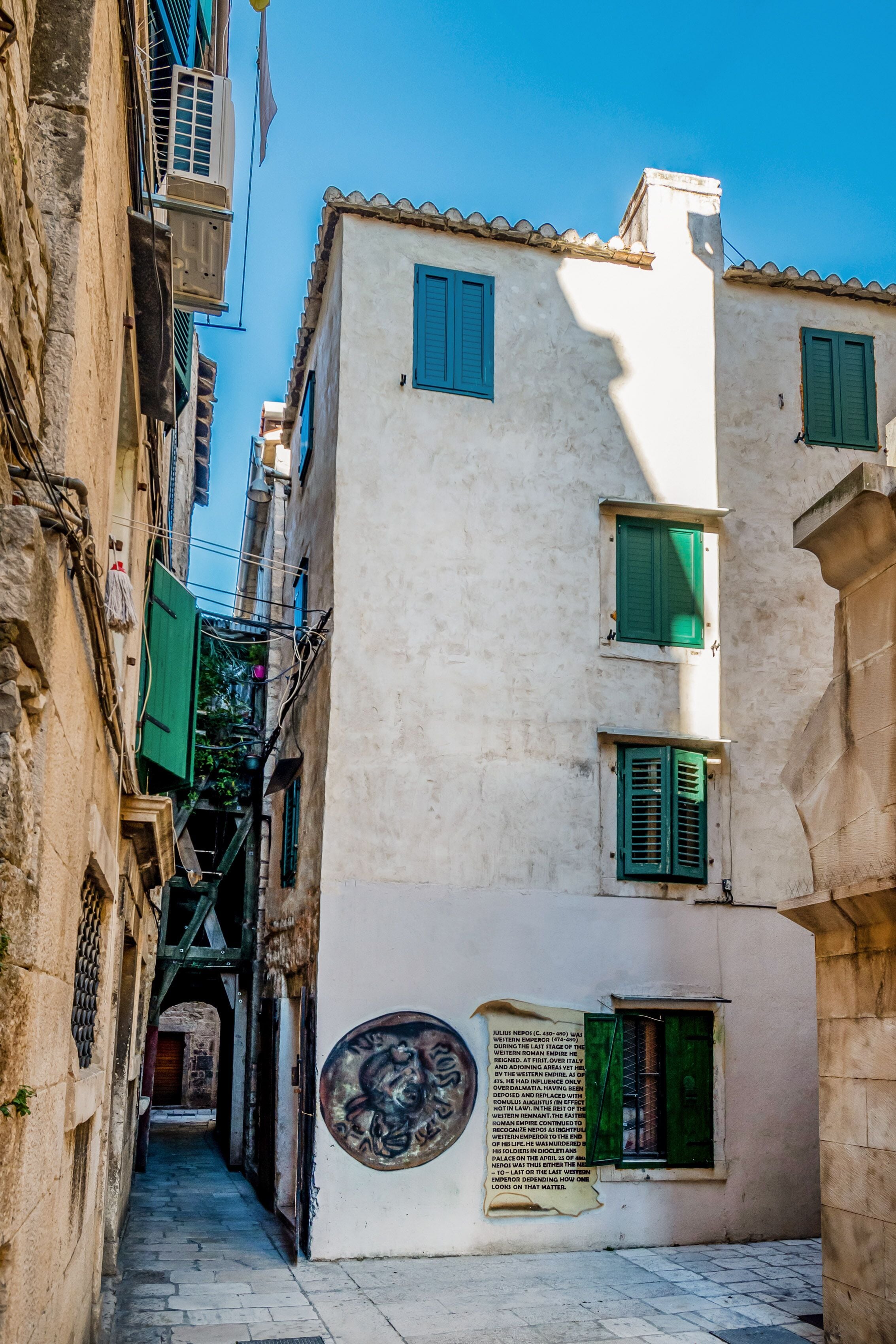 Un estrecho callejón flanqueado por edificios, con un mural y una pequeña ventana con contraventanas verdes.