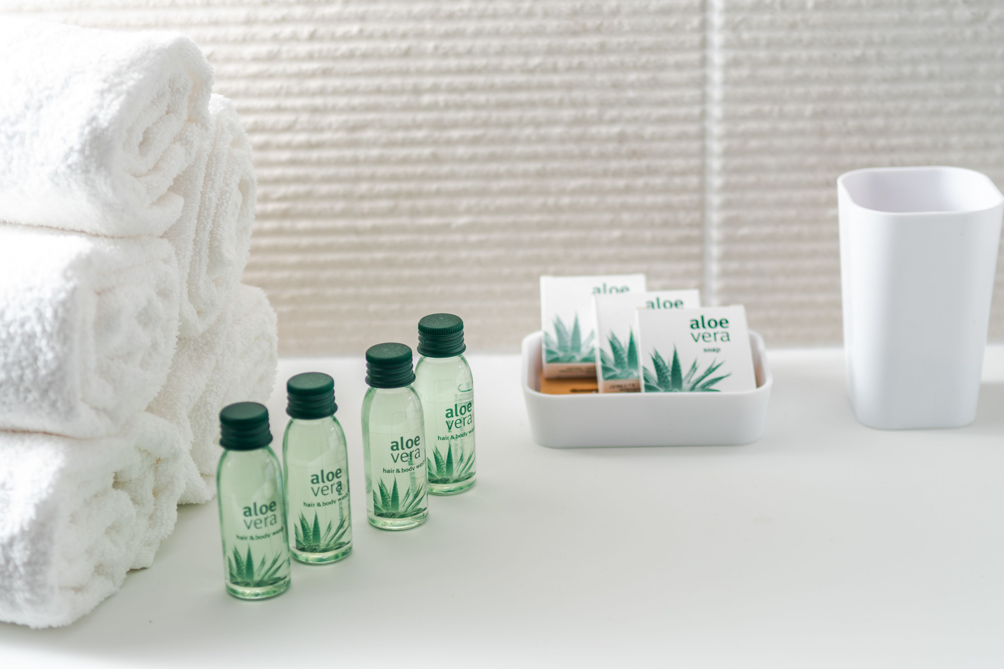 Un bagno con un mucchio di asciugamani bianchi, una tazza bianca e flaconi di detergente corpo e capelli all'aloe vera.