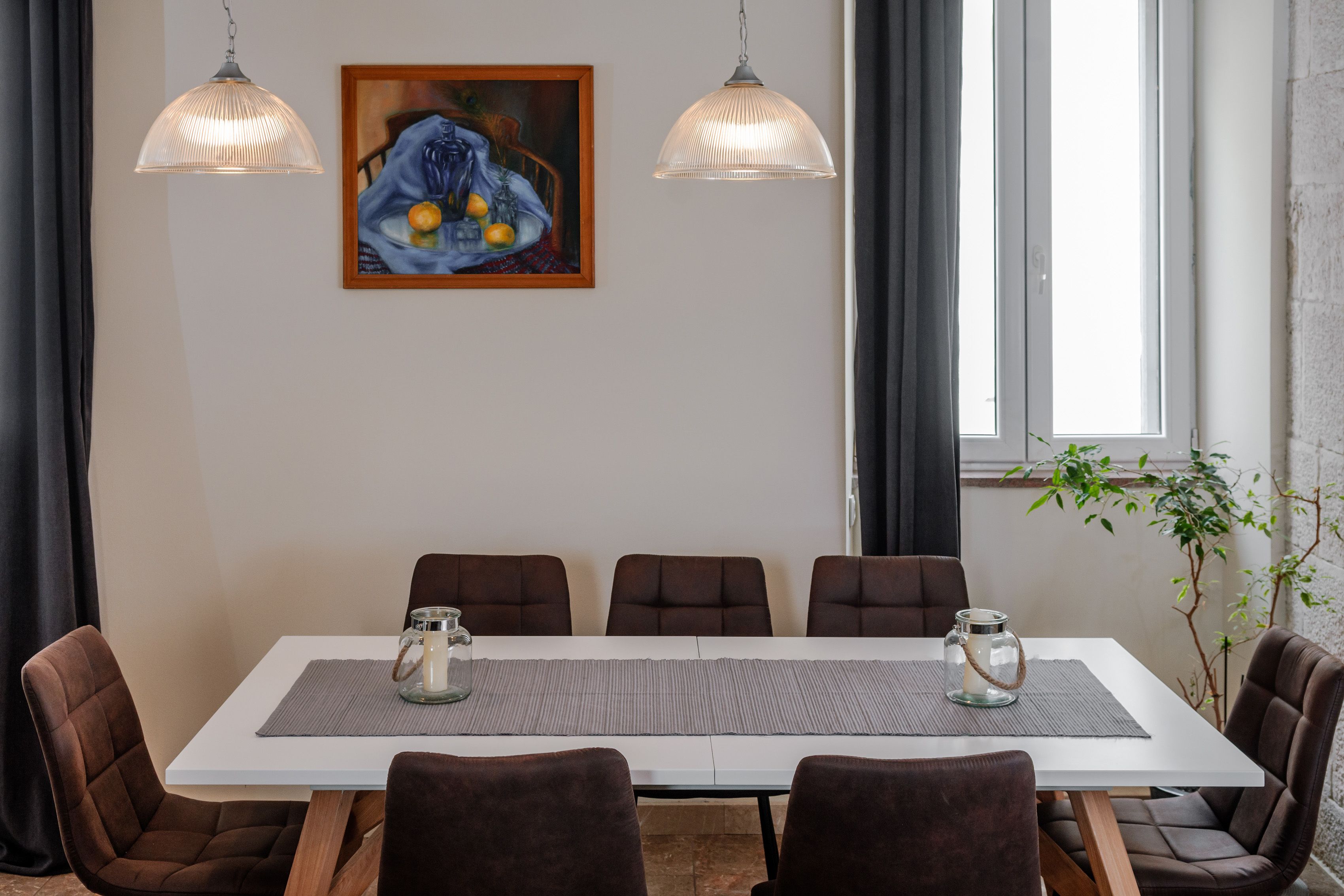 A dining area with a white table, six brown chairs, two pendant lights, and a painting on the wall.