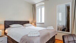 A neatly made bed with white linens and a brown headboard, a bedside table with a lamp, and a large mirror reflecting the room. - Bouganvillea Split Apartments (Split)
