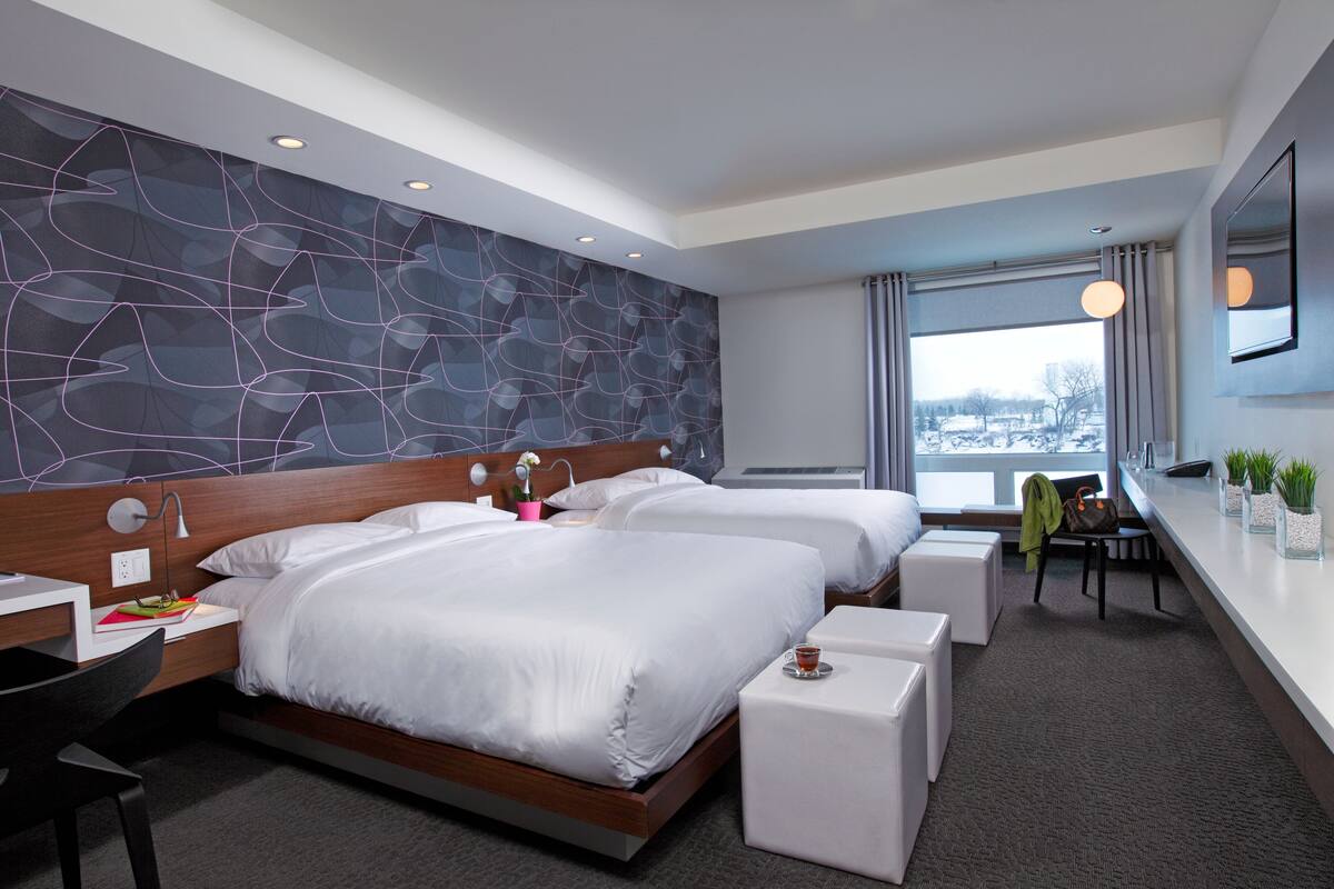 A modern hotel room with two beds, a desk, a chair, a small table, and a large window with a view.