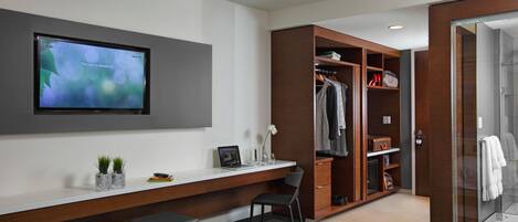 A modern hotel room with a bed, a desk with a laptop, a TV, a wardrobe, and a shower area.