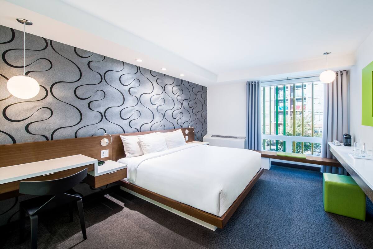 A modern hotel room with a large bed, a desk, and a chair, a small table, a green ottoman, and a view of the outside through a window.