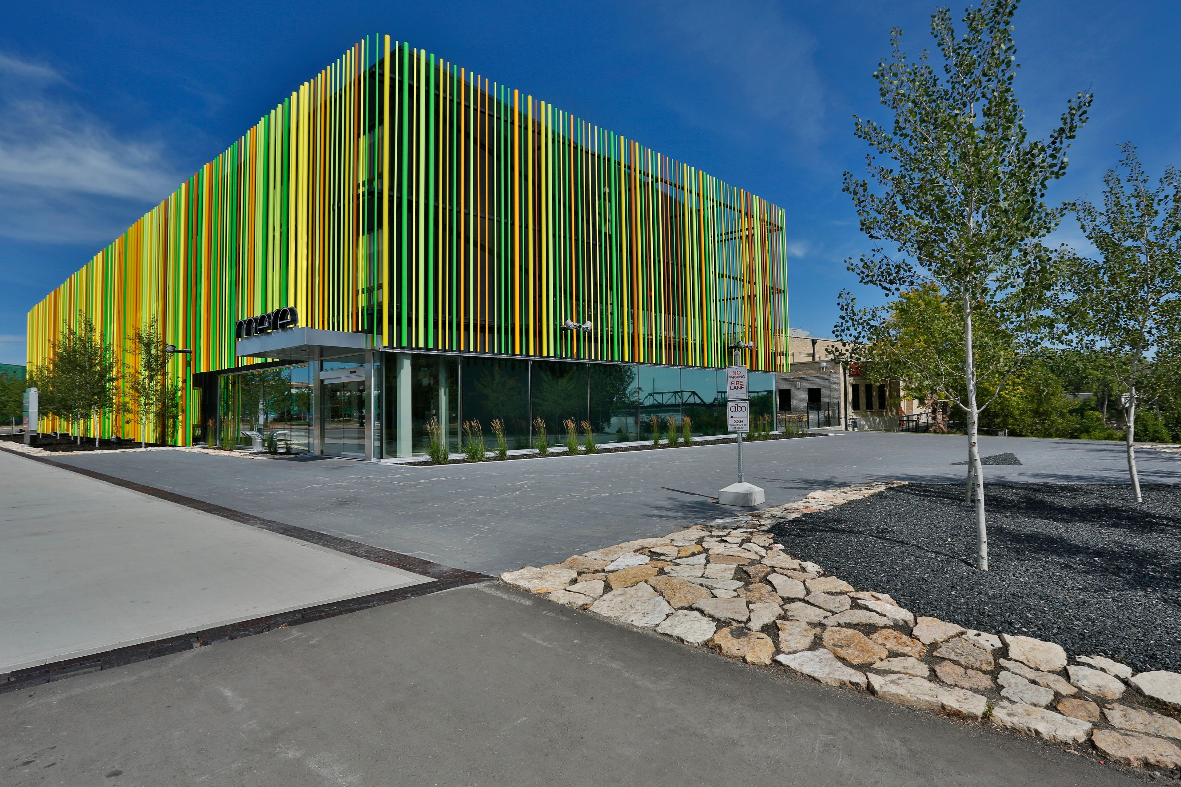 A modern building with vertical green and yellow stripes, a glass entrance, and a stone pathway with trees.