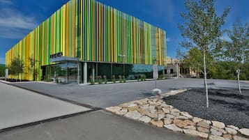 A modern building with vertical green and yellow stripes, a glass entrance, and a stone pathway with trees.