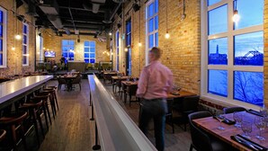 A restaurant with brick walls, large windows, and a long counter with stools. - Mere Hotel (Winnipeg)