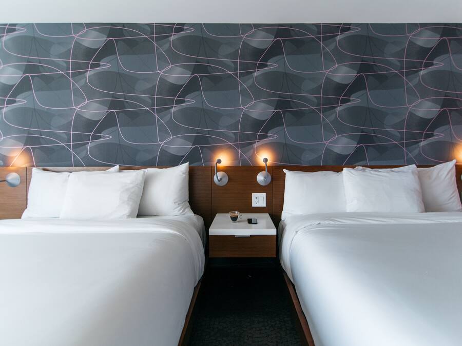 Habitación de hotel con dos camas, cabecera de madera, mesita de noche con lámpara y un diseño moderno y abstracto en la pared.