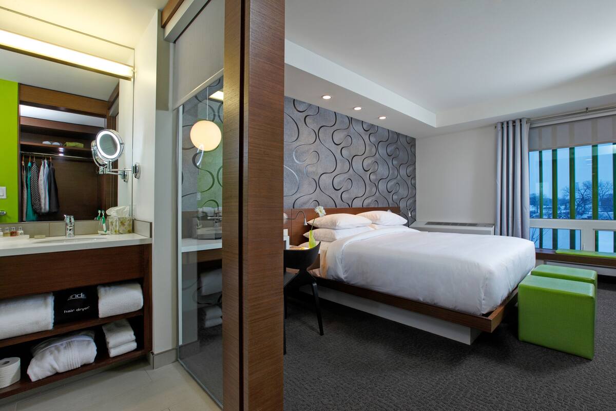 A hotel room with a bed, a vanity with a mirror, a green wall, and a window with a view.