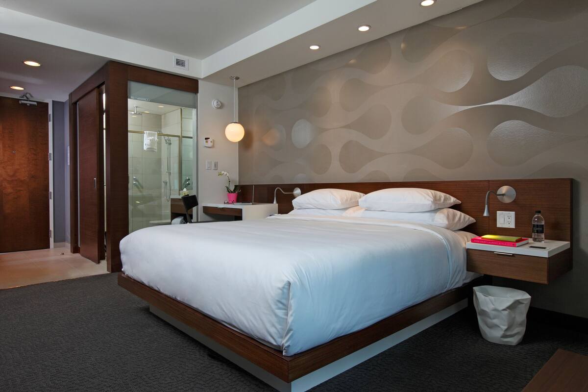 A modern hotel room with a large bed, a bedside table with a lamp, a nightstand with a bottle, and a trash bin.