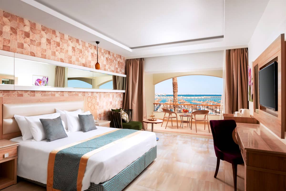 A hotel room with a large bed, a desk, a chair, a TV, and a balcony with a view of the sea.
