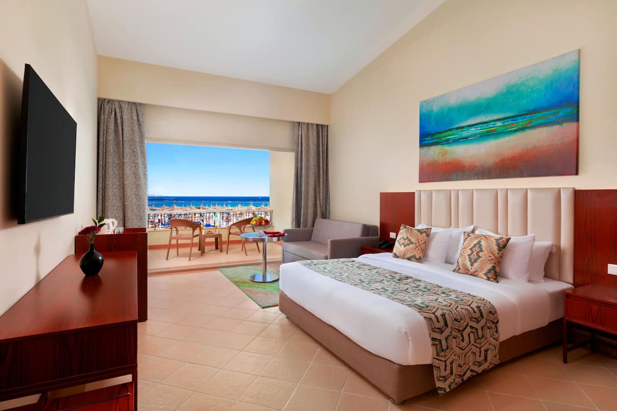 A hotel room with a large bed, a TV, a painting, a balcony with a view of the beach, and a dining area.