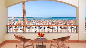 A balcony with wicker chairs, a small table with a fruit bowl, and a view of a beach with umbrellas and the sea.