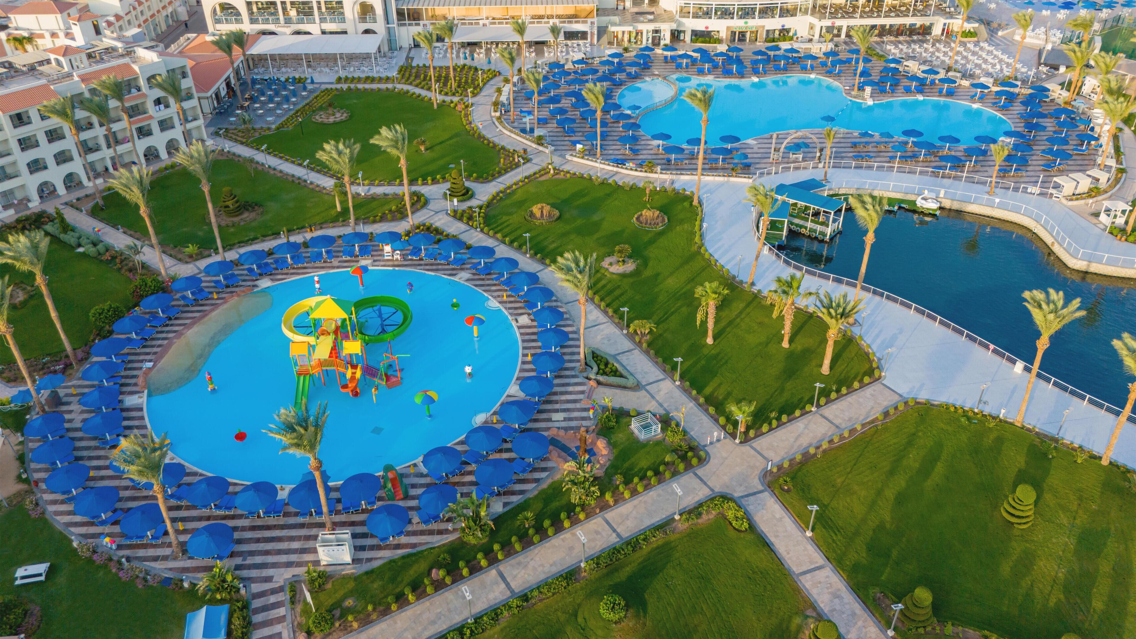 A large hotel complex with multiple swimming pools, palm trees, and a children's play area.