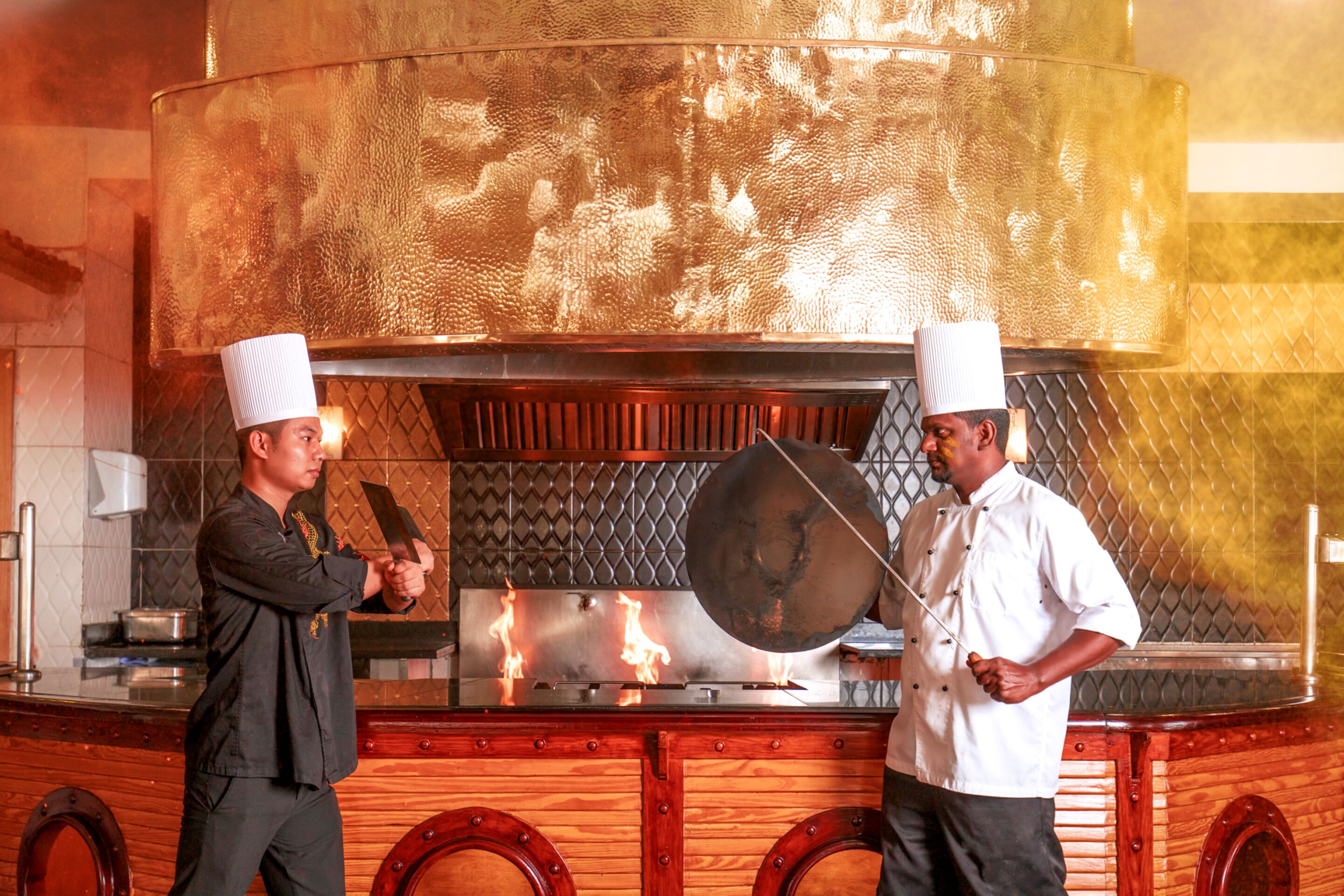 Deux chefs se trouvent dans une cuisine équipée d’un grand poêle décoré et d’un comptoir en bois poli.