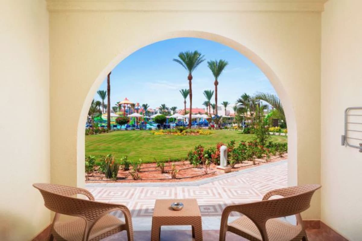 A room with a view through an archway to a pool area, including a lawn, palm trees, and a building with a red roof.