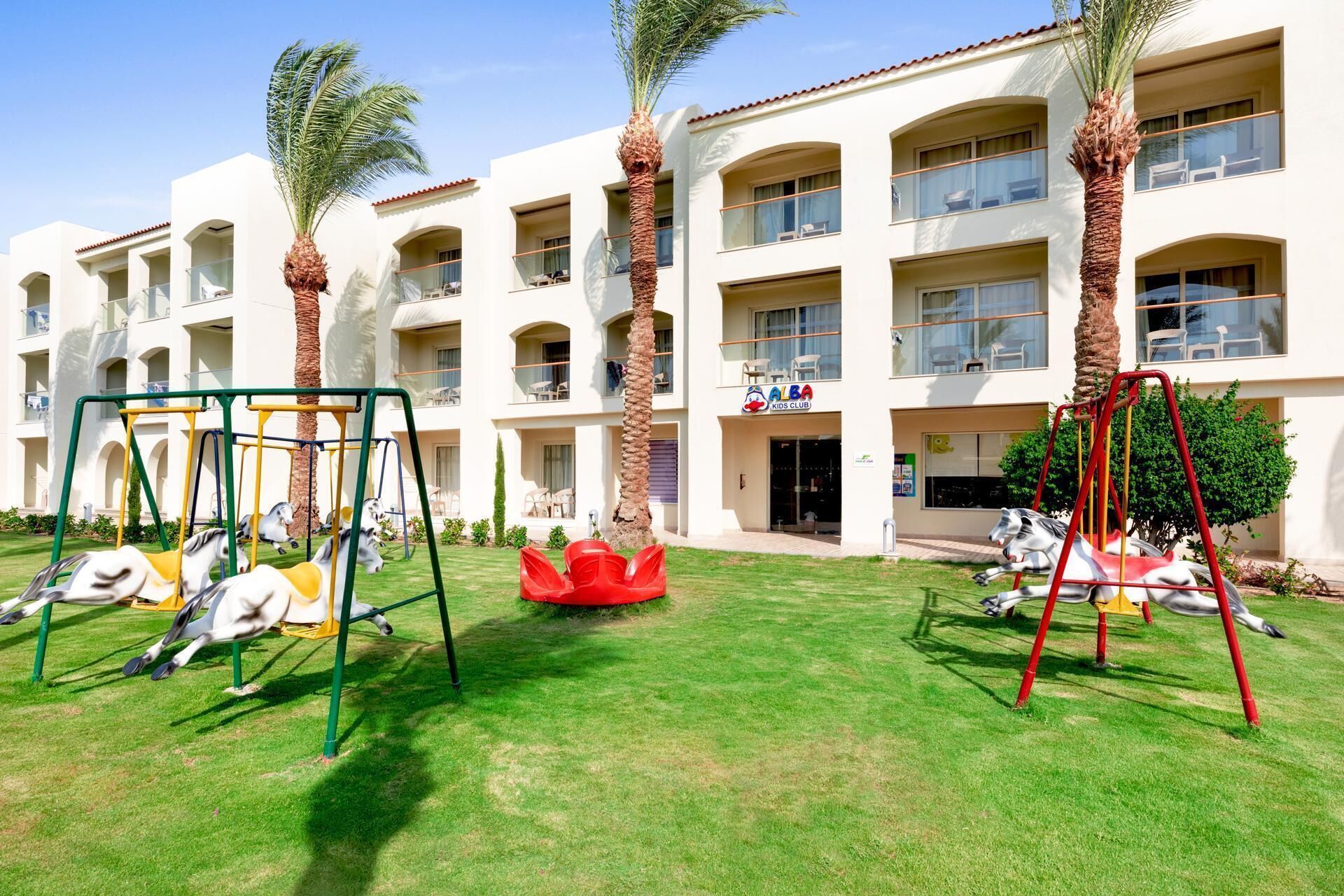 A children's play area with swings and a carousel, surrounded by a building with multiple balconies and a sign for a kids club.