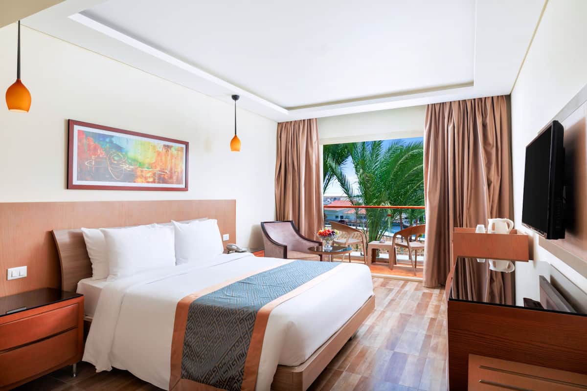 A hotel room with a large bed, a desk, a chair, a TV, and a balcony with a view of palm trees.