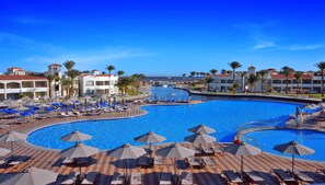 A resort pool area with multiple lounge chairs and umbrellas, surrounded by palm trees and buildings with red-tiled roofs.