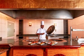 A chef in a professional kitchen with a large stove, a stainless steel countertop, and a variety of dishes displayed.