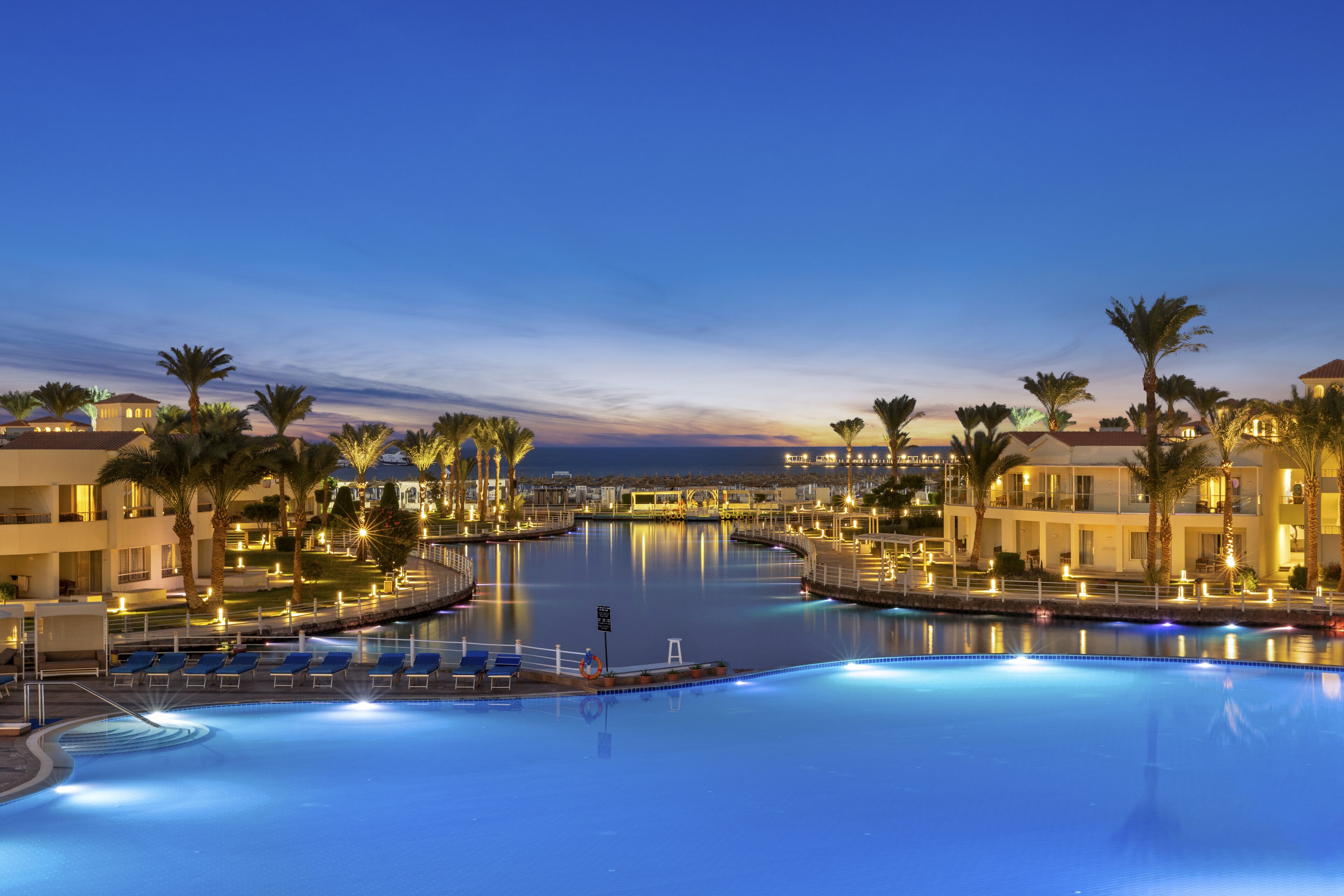 A swimming pool surrounded by palm trees and hotel buildings at dusk.