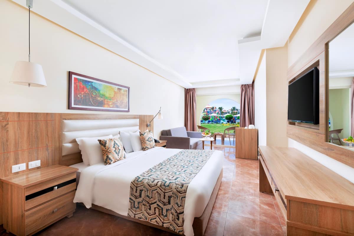A hotel room with a large bed, a TV, a desk, and a view of a garden.