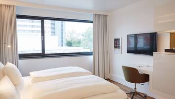Ein Hotelzimmer mit großem Fenster, einem Bett mit weißer Bettwäsche, einem Schreibtisch mit Stuhl und einem Flachbildfernseher.