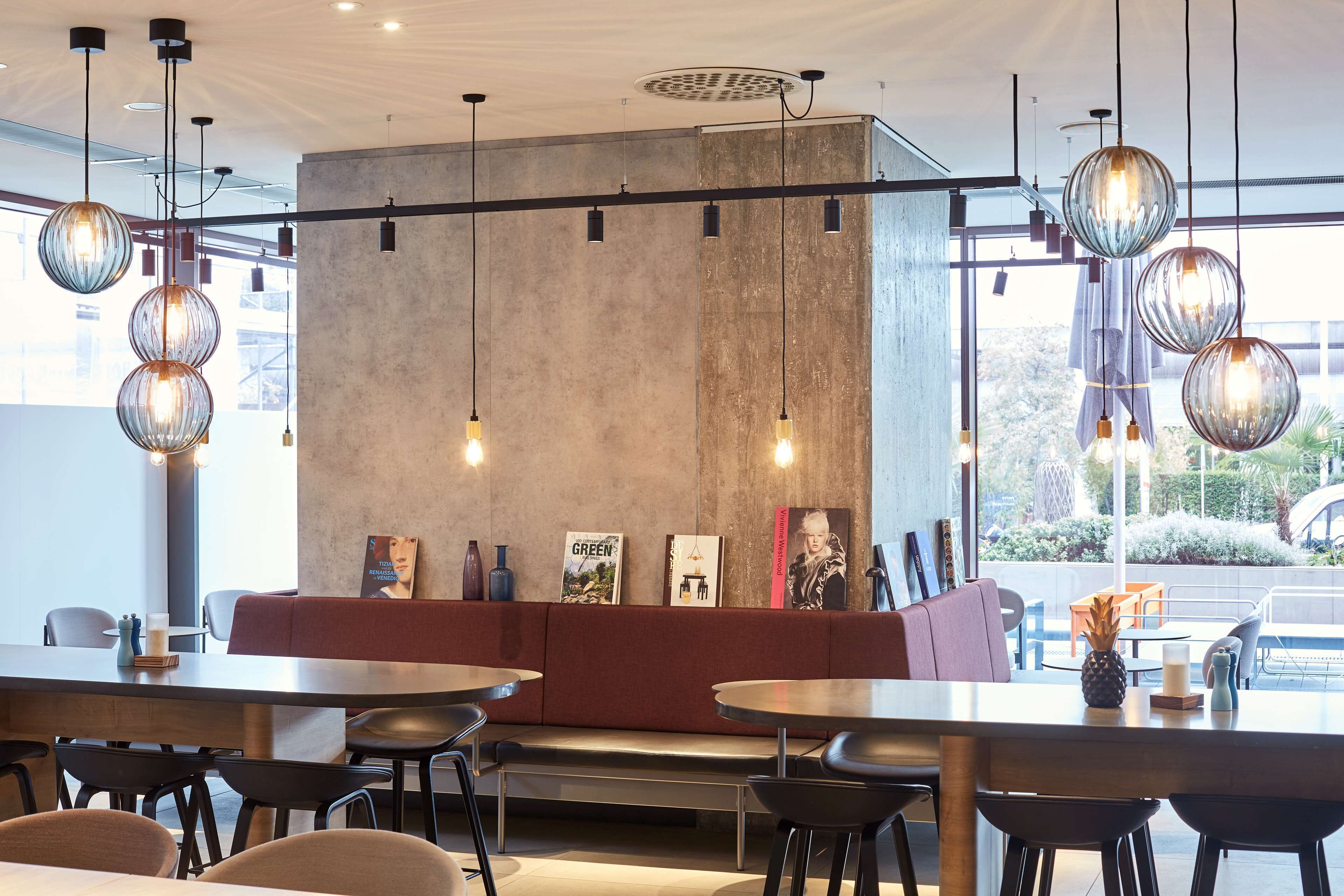 Interior moderno de restaurante con mesas redondas, un banco con cojines rojos, lámparas esféricas colgantes y una pared decorativa de hormigón.