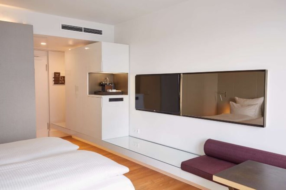 Una habitación de hotel moderna con cama, zona de estar, espejo grande y una kitchenette pequeña.