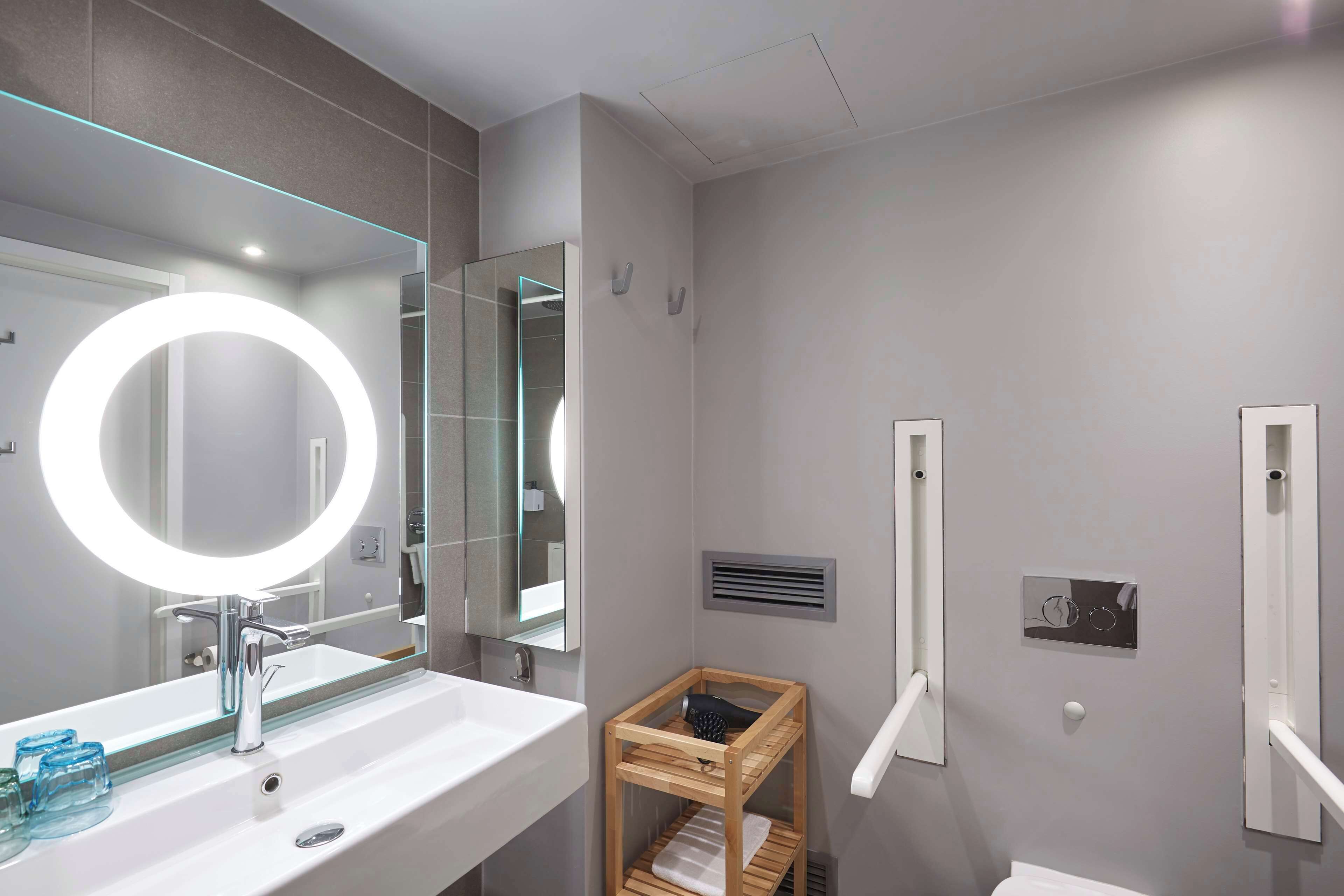 A modern bathroom with a large mirror, a round light, and a wooden shelf.
