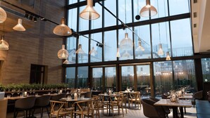 A modern restaurant with large windows, wooden tables, and hanging pendant lights.
