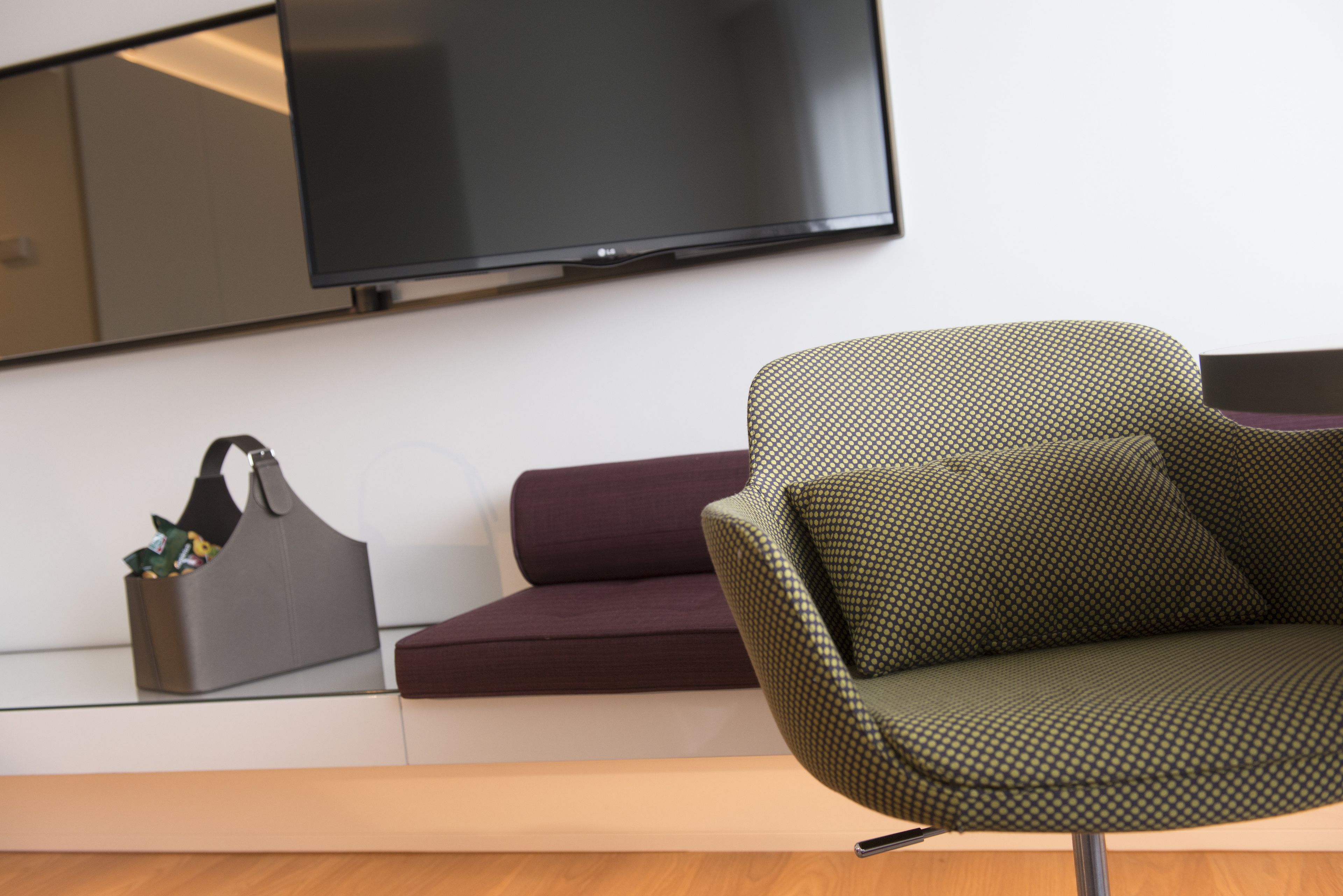 Una sala moderna con un televisor de pantalla plana montado en la pared, un bolso de cuero marrón oscuro sobre una mesa consola blanca y un sillón verde estampado.