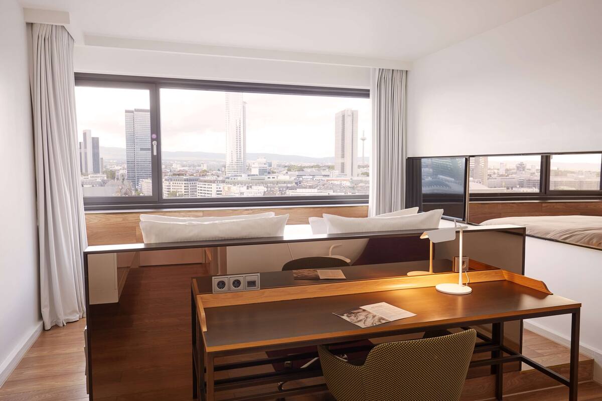 A hotel room with a large window, a desk, a chair, a bed, and a TV.