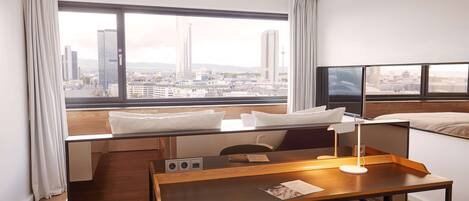 A hotel room with a large window, a desk, a chair, a bed, and a TV.