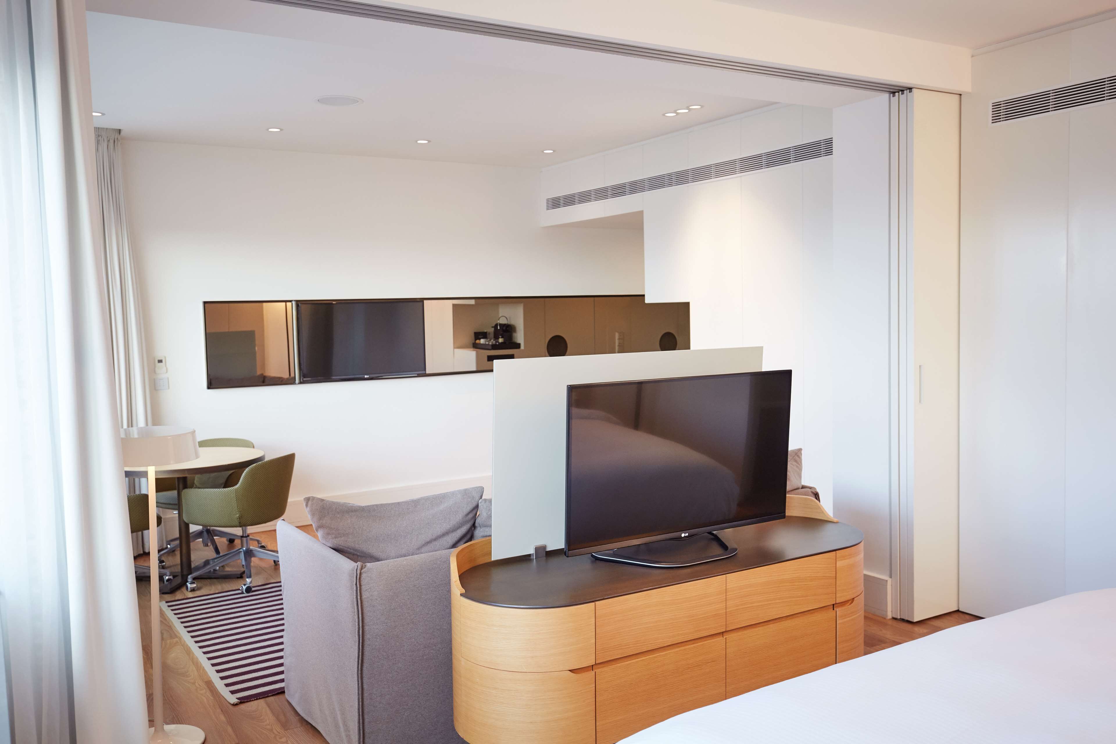 A modern hotel room with a flat-screen TV on a wooden console, a bed, a desk with a chair, and a dining area with chairs.