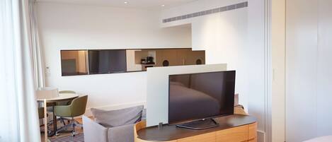 A modern hotel room with a flat-screen TV on a wooden console, a bed, a desk with a chair, and a dining area with chairs.