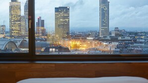 A cityscape view from a room with a wooden floor and a bed in the foreground.