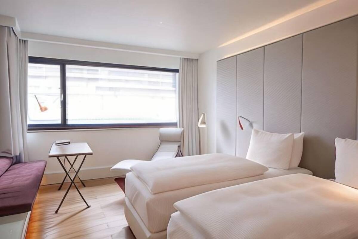 A hotel room with two beds, a small table, a chair, and a window with curtains.