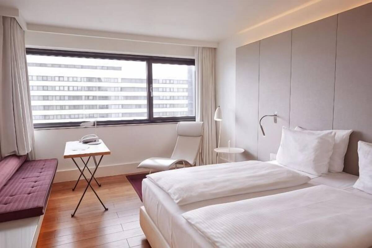 A hotel room with two beds, a small table, a chair, a desk, and a window with a city view.