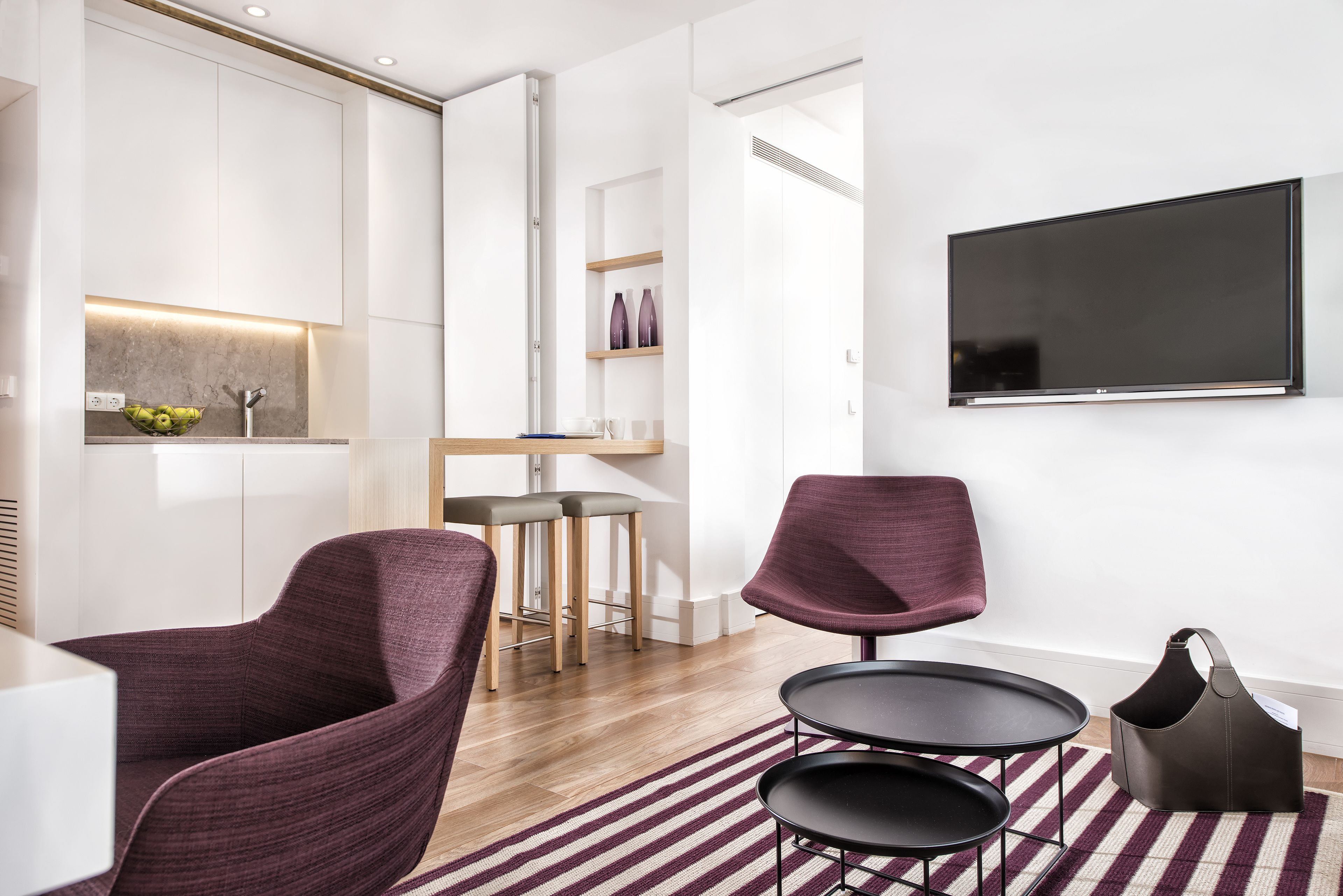 A modern living room with a flat-screen TV, two purple chairs, a small round table, and a dark brown bag on a striped rug.