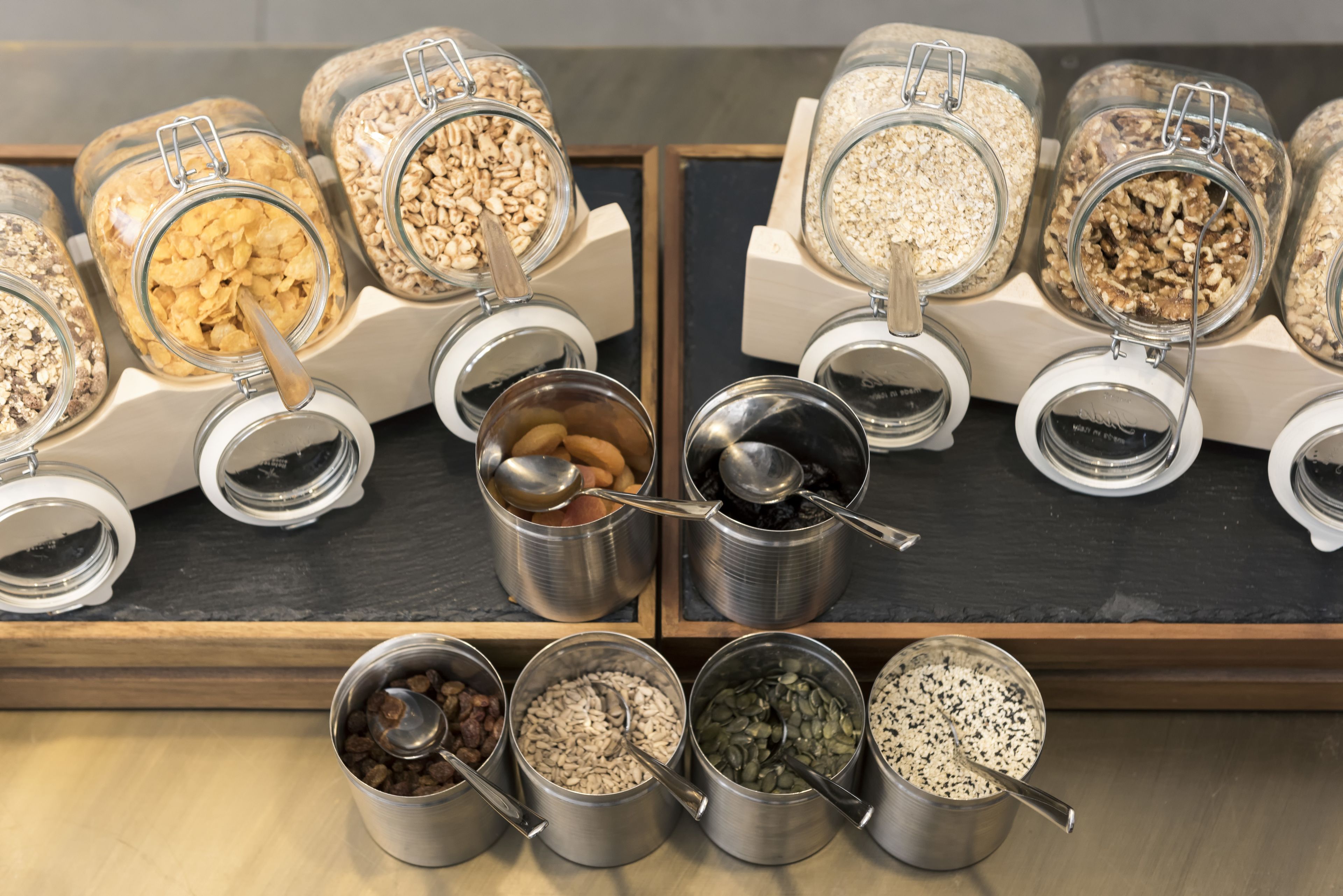 Une variété d’aliments, présentés dans des récipients en métal accompagnés de cuillères, disposés sur un comptoir en bois.