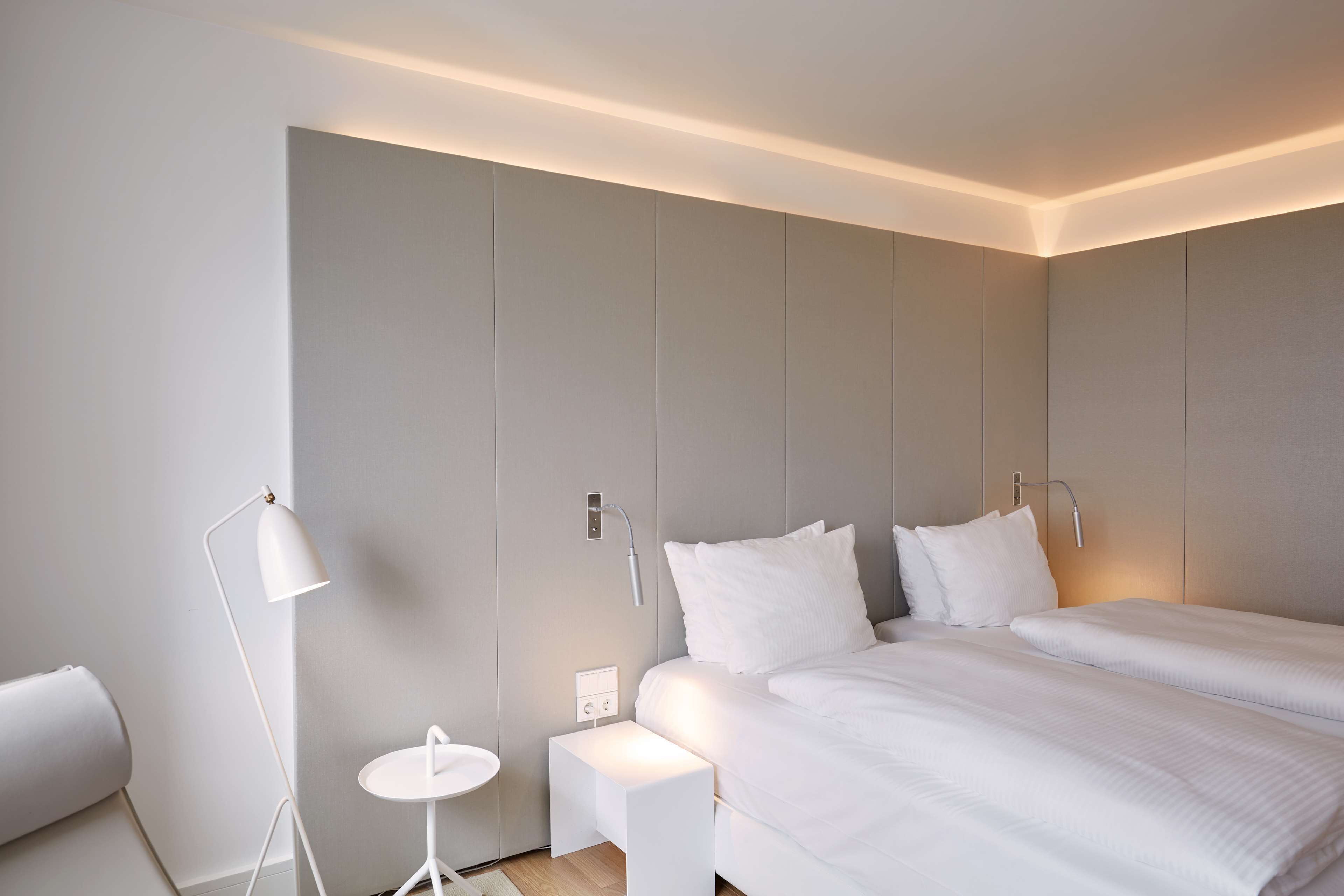 A modern hotel room with two beds, a bedside table, a floor lamp, and a small round table.