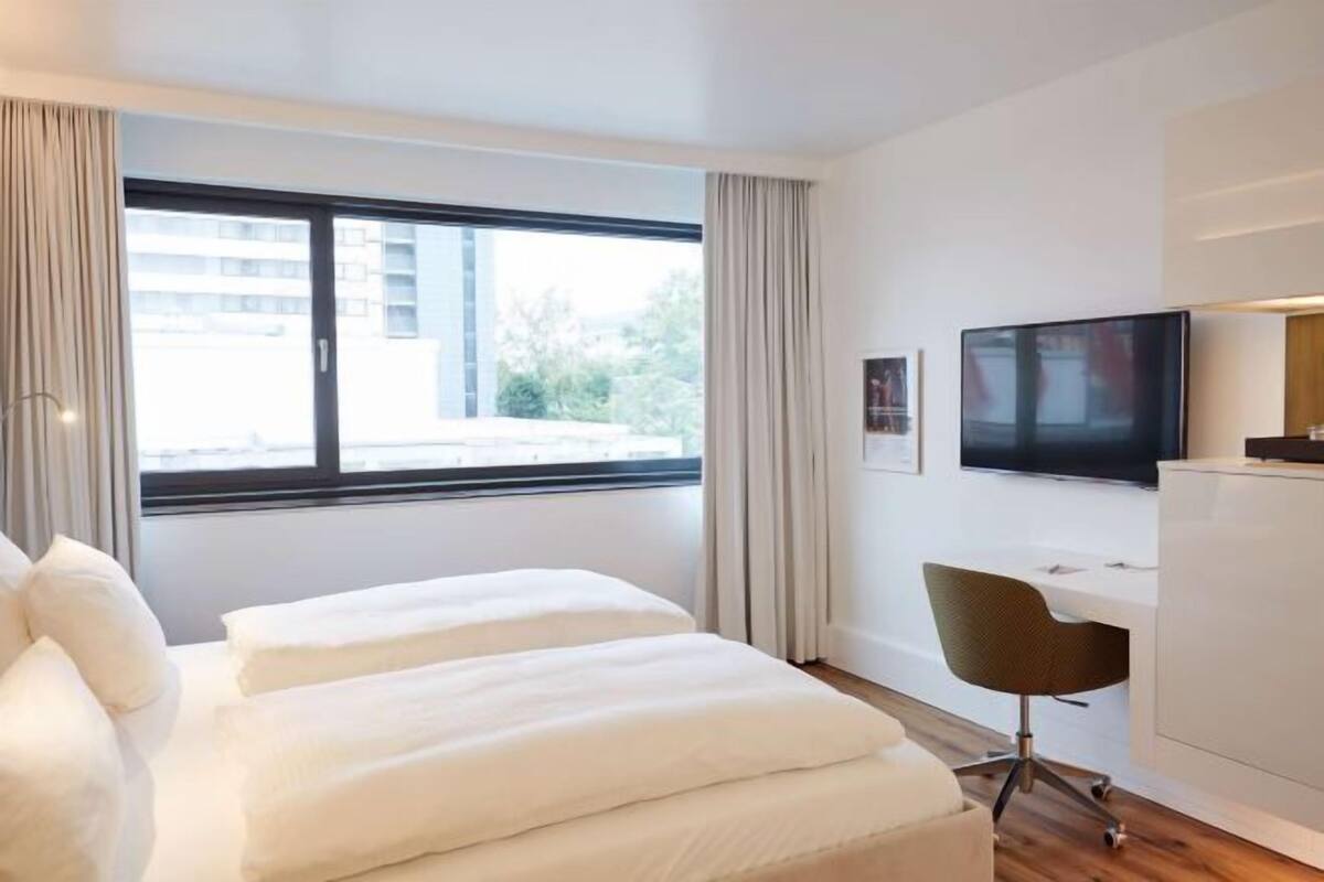 A modern hotel room with a large window, a bed, a desk, and a television.