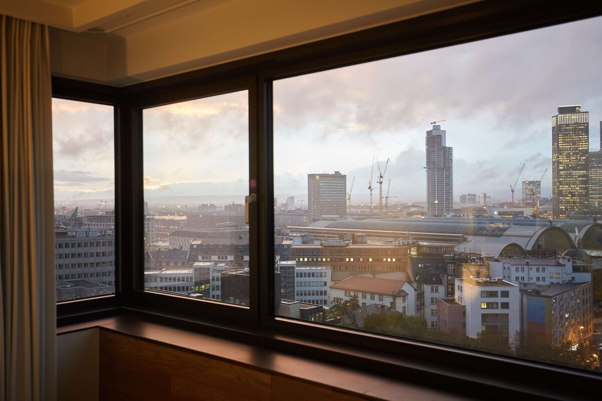 A view from a window with a cityscape, including buildings and cranes, during daylight.