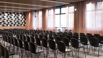 Un salón de conferencias con filas de sillas, un papel tapiz estampado y ventanas amplias con cortinas.