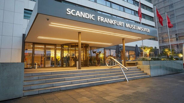 The exterior of the Scandic Frankfurt Museumufer hotel with a modern facade, large glass windows, and a staircase leading to the entrance.