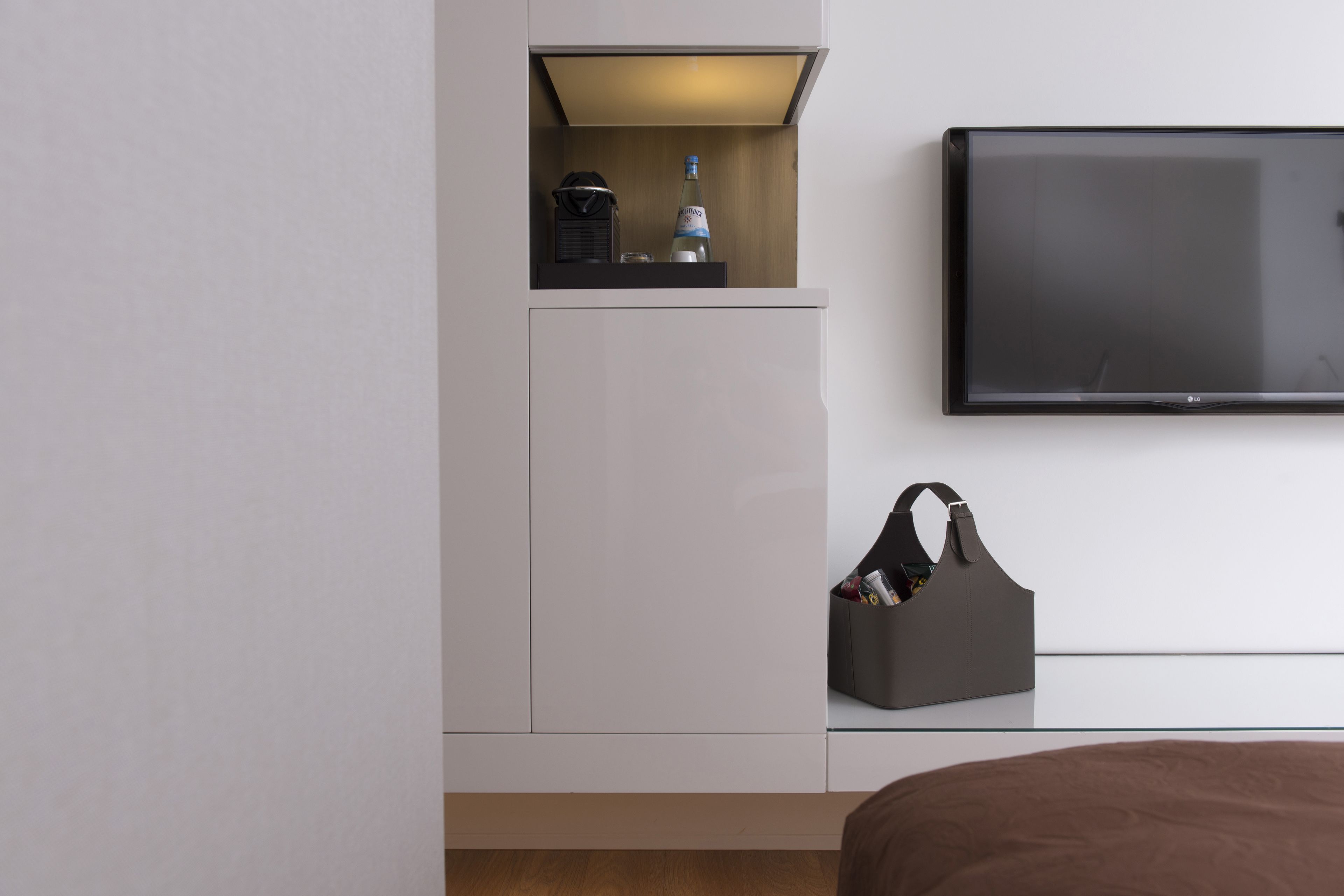 A modern hotel room with a bed, a TV mounted on the wall, a minibar, and a storage unit.