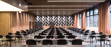 Sala de conferência com mesas e cadeiras dispostas em fileiras, parede com padrão geométrico e amplas janelas com cortinas.