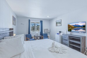 Une chambre d’hôtel avec un lit, deux chaises, une petite table, un bureau, une télévision et une fenêtre avec des rideaux.
