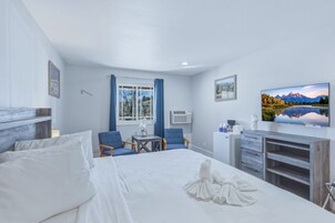 Habitación de hotel con una cama, dos sillas, una mesita, un escritorio, televisor y ventana con cortinas.
